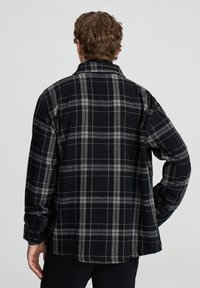 Mężczyzna z kręconymi brązowymi włosami stoi tyłem, ubrany w długą koszulę z czarno-szarym wzorem w kratę, z ręką na biodrze na jednolitym tle.