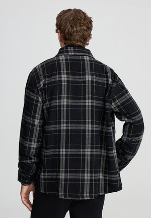 Uomo con capelli ricci castani che sta girato di spalle, indossa una camicia a maniche lunghe a scacchi nera e grigia, con una mano appoggiata all'anca su uno sfondo semplice.