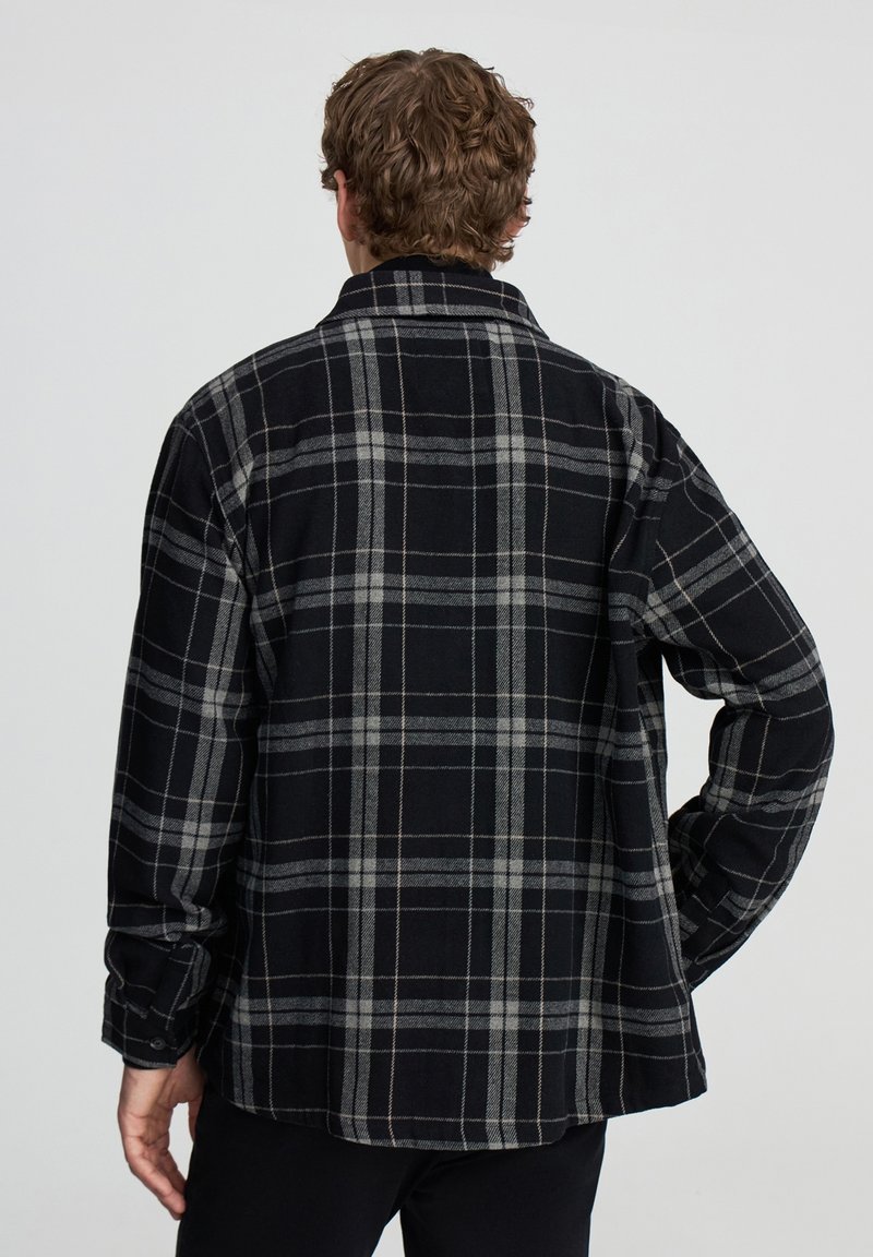 Mężczyzna z kręconymi brązowymi włosami stoi tyłem, ubrany w długą koszulę z czarno-szarym wzorem w kratę, z ręką na biodrze na jednolitym tle.