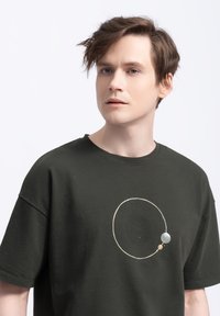 Donkergroen katoenen T-shirt met een cirkelvormig, minimalistisch graphic design met een kleine lichte grijze accenten, gepositioneerd in het midden van de voorkant.