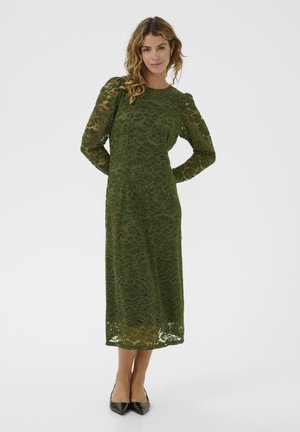 Woman standing with hands behind back wearing a long-sleeve, olive green lace midi dress and black pointed flats on a white background.