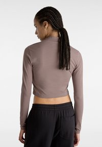 Maglietta corta a maniche lunghe in tessuto marrone chiaro, con collo alto e superficie liscia, abbinata a pantaloni felpati neri con dettagli visibili delle tasche.