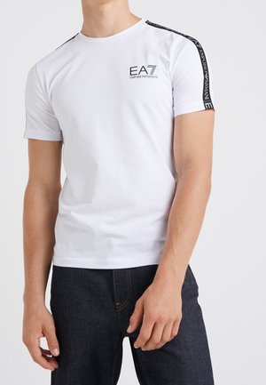 Mann trägt ein weißes kurzärmeliges EA7 Emporio Armani T-Shirt mit schwarzen Markenstreifen auf den Schultern und dunkle Jeans.