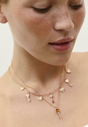 Podrobno posnetek ženskega vratu z dvema zlatima verigama, na katerih sta obeska v obliki srca in ključa, okrašena z majhnimi dragulji.