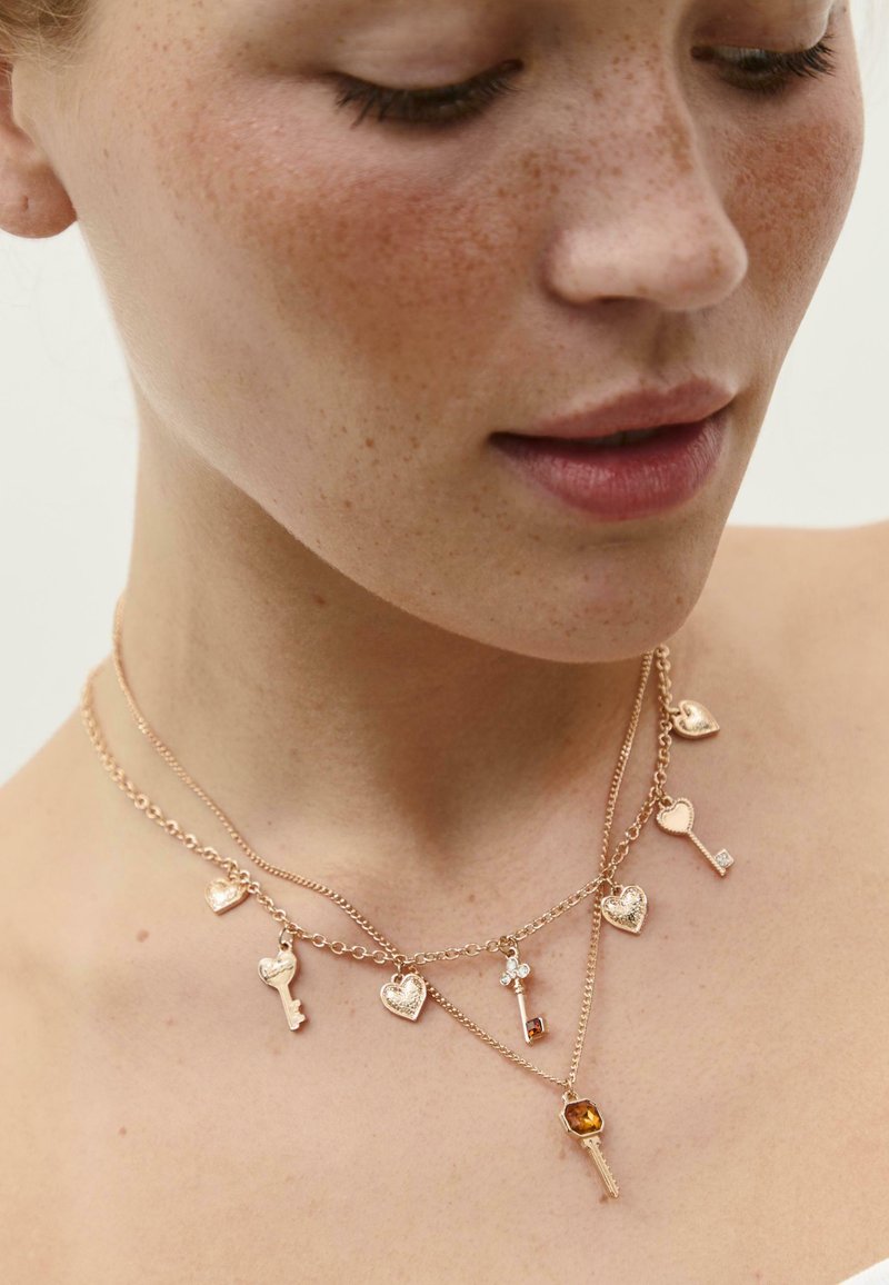 Uvećan prikaz ženskog vrata s dva zlatna lanca koja nose privjeske u obliku srca i ključa s malim gem dodacima.