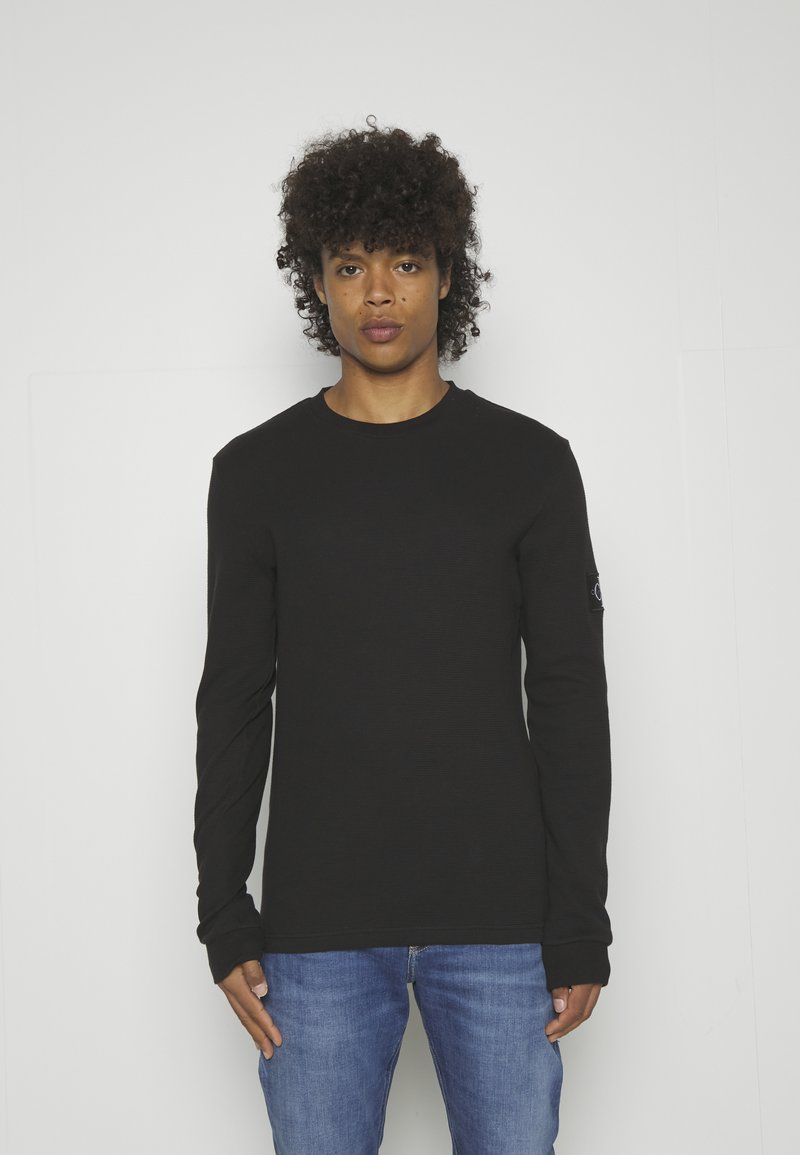 Jonge persoon met krullend haar, gekleed in een zwart shirt met lange mouwen en een blauwe spijkerbroek, staand tegen een effen witte achtergrond.