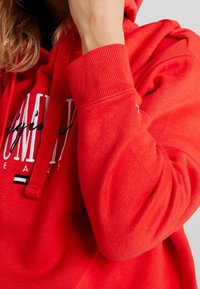 Person trägt einen leuchtend roten Hoodie mit teilweise sichtbarem weißen und schwarzen Sticktext und hält eine Hand nah am Gesicht.