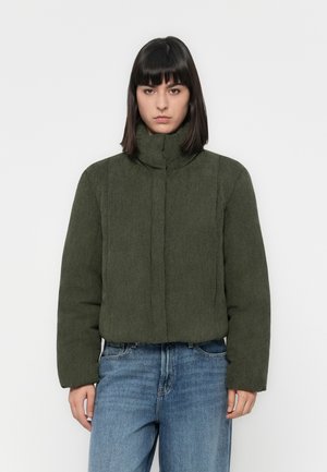 Femme à hair noir lisse portant une veste côtelée olive et un jean bleu, se tenant devant un fond blanc uni.
