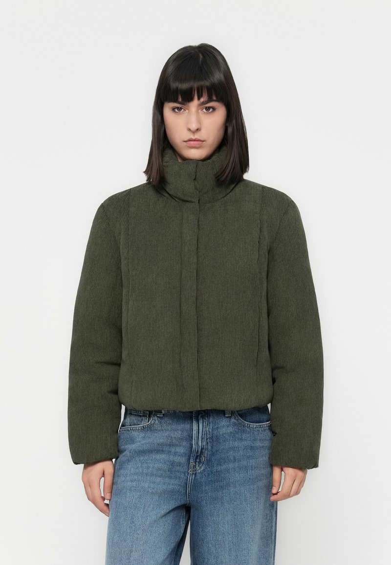 Vrouw met steil donker haar draagt een olijfgroene ribjack en blauwe jeans, staand tegen een effen witte achtergrond.