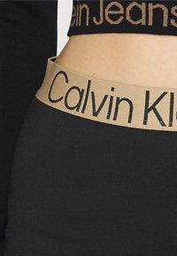 Svarta tajta leggings med en slät textur, innehållande en kontrasterande tan elastisk midjeband prytt med "Calvin Klein"-märke.