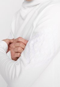 Top bianco a maniche lunghe con collo alto, caratterizzato da dettagli testurizzati sulle spalle e maniche aderenti. Tessuto liscio con vestibilità attillata.