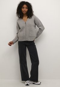 Cardigan grigio con zip, dotato di due tasche frontali, polsini a coste e una texture morbida, abbinato a jeans neri a gamba larga e scarpe sportive.