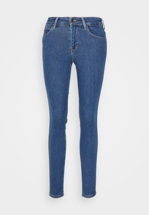 Jean skinny en denim bleu avec une texture lisse, cinq poches, passants de ceinture et fermeture éclair, présentant des coutures contrastées le long des coutures.