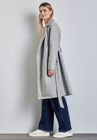 Szary wełniany płaszcz z długimi rękawami, z boku związany i o swobodnym kroju, połączony z szerokimi, ciemnoniebieskimi dżinsami oraz białymi trampkami.