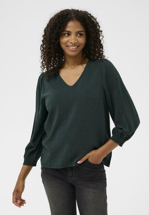 Blouse vert foncé avec un décolleté en V et un tissu texturé. Présente des manches bouffantes avec des poignets froncés. Associée à un jean foncé.
