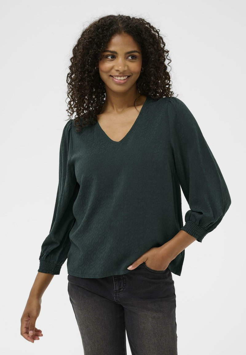 Donkergroene blouse met een V-hals en gestructureerde stof. Heeft pofmouwen met gathers bij de manchetten. Gecombineerd met donkere jeans.
