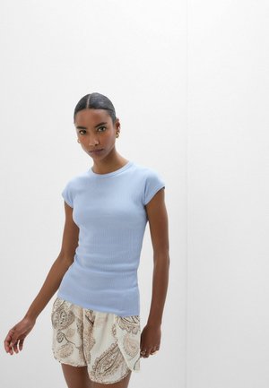 Kvinde med mørkt hår iført en lyseblå tætsiddende top med korte ærmer og cremefarvede shorts med paisley-mønster mod en ensfarvet hvid baggrund.