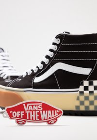 Čern�é vysoké tenisky se bílými tkaničkami a charakteristickým bílým pruhem Vans. Mají kostkovaný akcent a červený štítek s logem Vans.