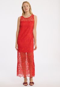 Rotes Spitzenkleid mit ärmellosem Design, aufwendigen floralen Mustern, einem transparenten unteren Abschnitt und einem runden Ausschnitt. Dazu offene Schuhe.