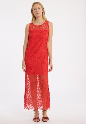 Rotes Spitzenkleid mit ärmellosem Design, aufwendigen floralen Mustern, einem transparenten unteren Abschnitt und einem runden Ausschnitt. Dazu offene Schuhe.