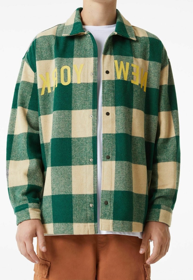 Chaqueta de cuadros verde y beige con "NUEVA YORK" en texto amarillo. Cuenta con cuello, mangas largas y cierre de botones a presión.