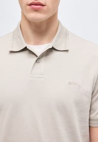 Homme portant un polo beige BOSS avec un col à boutons et un sous-pull blanc visible au niveau du cou.