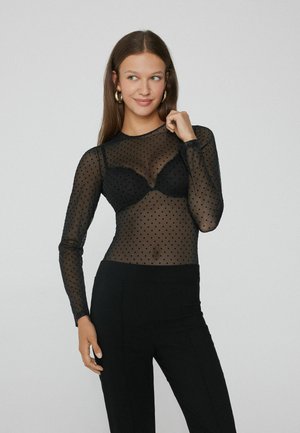 Schwarzes Mesh-Langarmoberteil mit Punkte-Muster, transparenter Textur und rundem Ausschnitt, kombiniert mit einem schwarzen BH und taillierten schwarzen Hosen.