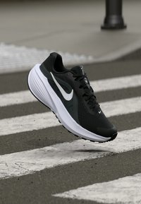 Μαύρο αθλητικό παπούτσι με ελαφρύ πλέγμα στην επάνω πλευρά, λευκές λεπτομέρειες και υφασμένη σόλα; διαθέτει κορδόνια και μια επιβλητική λογότυπο στο πλάι.