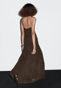 Woman with curly hair wearing a dark brown sleeveless maxi dress, silver bracelets, and black open-toe heels, walking on a black floor.