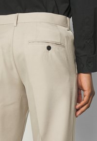 Pantaloni beige su misura con una texture liscia, dotati di una tasca posteriore a filetto e un bottone nero. Indossati con una camicia nera.