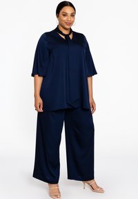 Marineblauwe two-piece outfit met een losse top met strik-hals en wijde broekspijpen, gemaakt van een zachte stof met een gedrapeerd ontwerp.