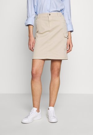 Kvinde iført beige cargo-nederdel til knæet, lyseblå langærmet skjorte og hvide sneakers, stående foran en ensfarvet baggrund.