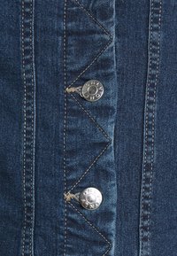 Detalj av denimjacka med blått tyg, silverfärgade tryckknappar, sicksackstygn och kontrasterande ljusbrunt tråd längs sömmarna.