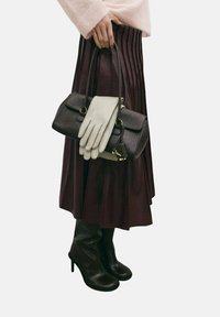 Bolso de cuero marrón con detalles dorados, guantes grises, falda plisada marrón oscuro y botas negras de tacón hasta la rodilla. Se aprecian materiales de textura suave.