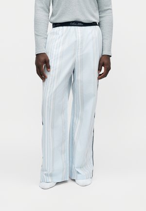 Personne portant un pantalon blanc rayé ample avec une ceinture Calvin Klein noire visible et un haut gris à manches longues, debout sur fond blanc.
