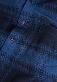Camicia in flanella a quadri blu e neri con chiusura a bottoni, taschino sul petto e una texture morbida caratterizzata da motivi orizzontali e verticali.