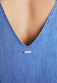 Blå denim topp med bred V-hals, med et lite sølvmerke som står "Pepe Jeans." Glatt tekstur med subtile sømdetaljer.