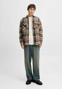 Camicia di flanella a quadri marrone e nera con due tasche sul petto, maglione crema sotto, pantaloni in denim chiaro larghi e scarpe chunky nere.