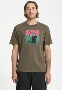T-shirt en coton vert olive avec un design graphique coloré représentant un soleil souriant et une silhouette en manteau noir. Manches courtes, coupe décontractée.