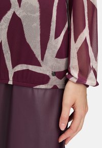 Dłoń obok spódnicy z gładkiego fioletowego materiału skórzanego oraz zwiewnej bluzki z długimi rękawami w abstrakcyjnym wzorze fioletowo-beżowym.