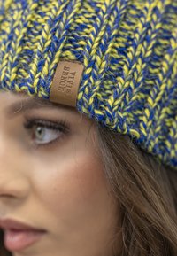Berretto lavorato a maglia in blu e giallo, con un motivo a costine e un'etichetta in pelle marrone con la scritta "VIVI SENCE" in rilievo. Materiale morbido e testurizzato.