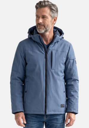 Mann mittleren Alters mit grauem Haar und Bart, der eine blaue Kapuzenjacke mit Reißverschluss und blaue Jeans trägt und leicht nach links schaut.
