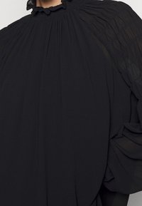 Blusa nera con colletto alto a volant, design plissettato e maniche a palloncino textured. Realizzata in un tessuto leggero e fluido.