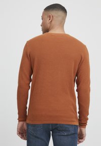 Pulover lung din tricot waffle, de culoare maro, cu guler rotund, având manșete și tiv ribbate, purtat cu blugi albaștri.