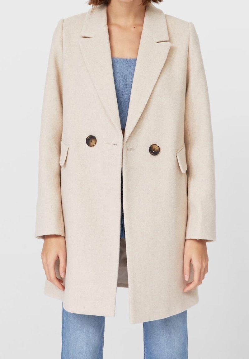 Abrigo beige de mezcla de lana con diseño de doble botonadura, solapas con muesca, dos bolsillos delanteros y botones redondos oscuros, llevado sobre una blusa azul.