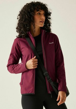 Kobieta o kręconych włosach, ubrana w bordową bluzę na zamek na czarnym topie, nosząca czarną torbę na ramię, patrząca w bok.