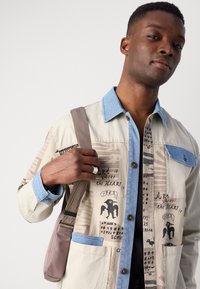 Beige Multi-Taschen-Jacke mit Denim-Kragen und blauen Akzenten, verziert mit abstrakten Mustern und Text, getragen mit einer braunen Umhängetasche.