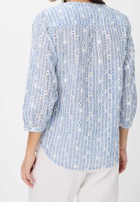 Blouse bleu clair avec broderie blanche, présentant un motif floral, coupe ample et manches longues légèrement bouffantes. Design avec ourlet arrondi.