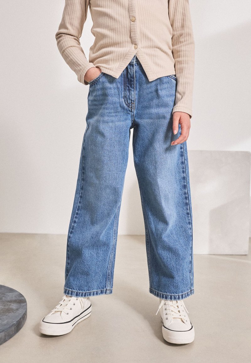 Persoon draagt een beige geribd shirt met knopen, wijde blauwe jeans en witte vetersneakers, staand met de handen in de zakken.