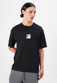 Black cotton t-shirt with a crew neckline and short sleeves, featuring a small white graphic logo centered on the chest.